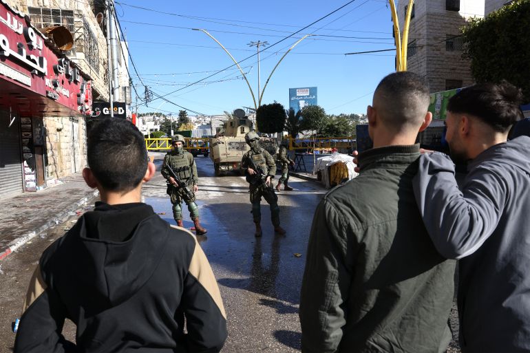afp_69711f9c5ccd-1769021340 Palestinians watch patrolling Israeli soldiers as the military closes part of the H2 southern sector of the West Bank city of Hebron, announcing a curfew as they search for weapons and Palestinians on their wanted list, in the Israeli occupied West Bank on January 19, 2026.