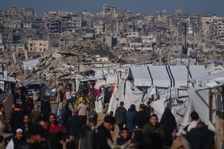 ap_69550a548cd3b-1767180884 فلسطينيون يسيرون قرب مخيم خيام في غزة أمام مبانٍ مدمرة بفعل العمليات العسكرية الإسرائيلية الجوية والبرية، في مشهد يجسد دمار الأحياء السكنية واستمرار معاناة النازحين.