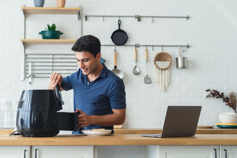 fg76gy-1769600952 Happy handsome man cooking healthy food with recipe by Air Fryer machine and using laptop computer in the modern kitchen at home on sunny day. Watching or Learning with Technology connection. by edward_indy @ ادوبي ستوك