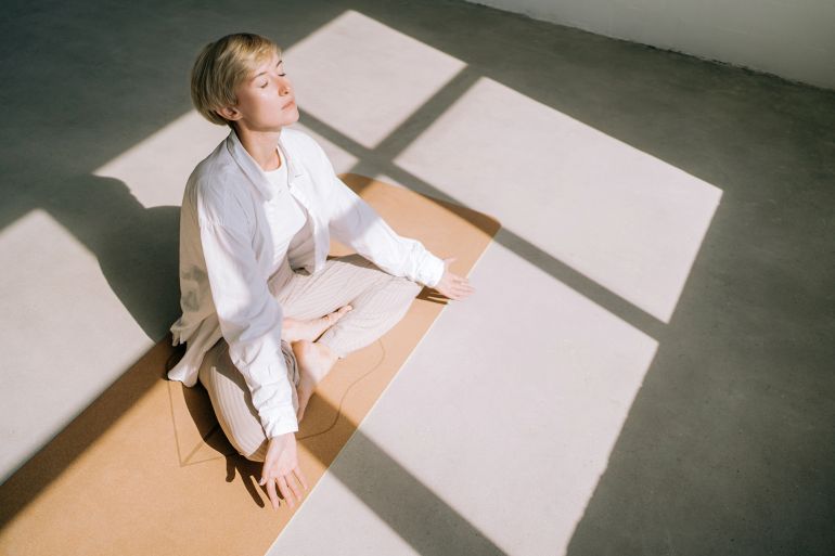 Beautiful authentic woman with short blond hair is meditating sitting in lotus position on yoga mat in front of a window. She is wearing a light-colored casual clothing. Concept of relaxation exercises