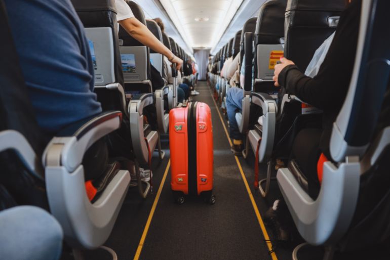 Interior of airplane with people sitting on seats. Passengers with suitcase in aisle looking for seat during flight.