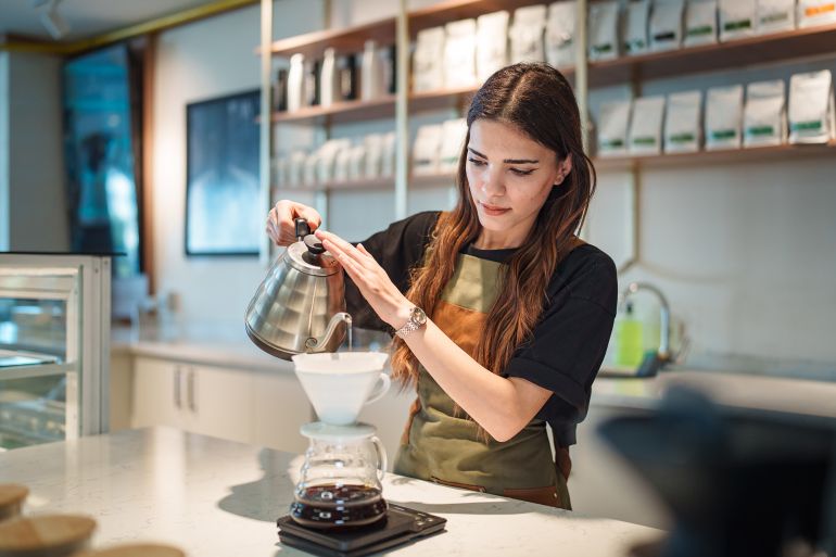 A female barista is carefully brewing pour-over coffee using a V60 dripper in a specialty café. The image captures the artisanal preparation process, emphasizing attention to detail, craftsmanship, and third-wave coffee culture. Ideal for themes such as slow coffee movement, manual brewing, café lifestyle, specialty coffee, and barista skills.