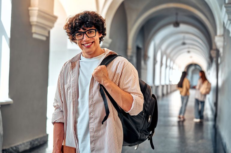 Handsome young confident college student. Smiling male student with backpack and books walking on campus indoors. Young man ready for classes or studying. Confident and happy college student. ; Shutterstock ID 2507996177; purchase_order: aj; job: ; client: ; other: