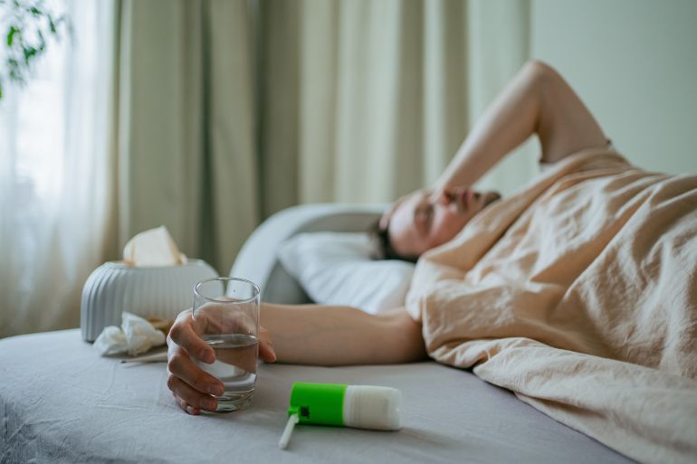 Sick man lying in bed at home, battling cold or flu symptoms, holding a glass of water while tissues and throat spray rest on the bedsheets in the foreground; Shutterstock ID 2675404423; purchase_order: aj; job: ; client: ; other:
