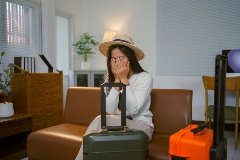 Asian woman is crying and covering her face with her hands while sitting on sofa with luggage in living room, travel problems concept