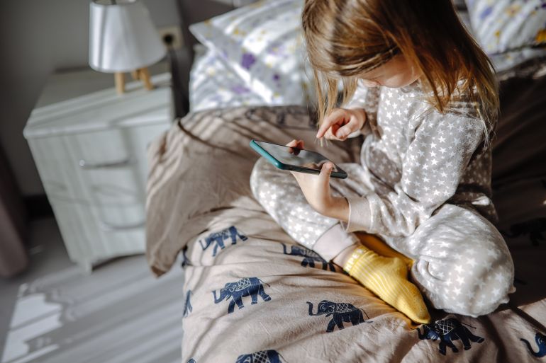 76tgh-1770619800 Child in pajamas scrolling on a smartphone while sitting on the bed in the morning, screen time and technology habits.