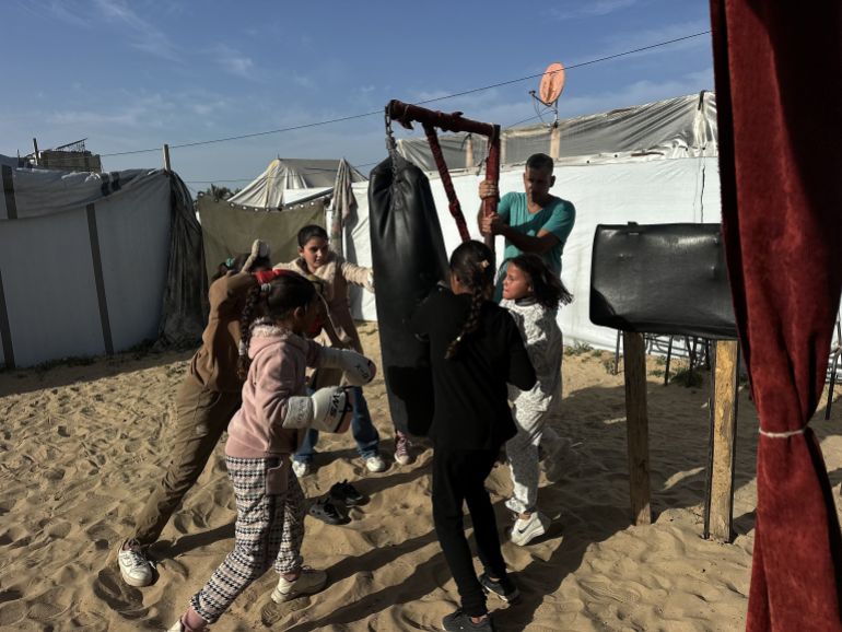 بين خيام النازحين في منطقة المواصي غربي مدينة خان يونس جنوبي قطاع غزة، تتدرب مجموعة من الفتيات الفلسطينيات على الملاكمة في إطار سعيهن للدفاع عن النفس وتفريغ الطاقة السلبية التي ولدتها حرب الإبادة الإسرائيلية على مدار عامين. الفتيات وجمعيهن دون سن السادسة عشرة، بدأن التدريبات خلال أشهر الإبادة، إذ تحولت هذه الرياضة المعروفة باسم "الفن النبيل" إلى وسيلة للصمود أمام الغارات والهجمات الإسرائيلية التي كانت تبث الرعب في نفوسهن. كما بدأت التدريبات بإمكانات بدائية وبسيطة مثل استبدال كيس الملاكمة بآخر من الرمال، وبلا قفازات وفرشات حماية، لكن في فترة لاحقة تم توفير أنواع وكميات محدودة من الأدوات الرياضية اللازمة. الكابتن أسامة أيوب، يقول للأناضول، إن اختيار هذا المكان بين خيام النزوح لإجراء هذه التدريبات كان قسريا، بعد أن أحرقت إسرائيل النادي المخصص في مدينة غزة خلال أشهر الإبادة. ( Doaa Albaz - وكالة الأناضول )