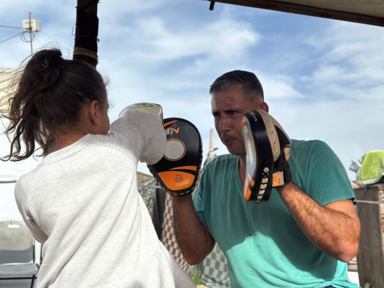 بين خيام النازحين في منطقة المواصي غربي مدينة خان يونس جنوبي قطاع غزة، تتدرب مجموعة من الفتيات الفلسطينيات على الملاكمة في إطار سعيهن للدفاع عن النفس وتفريغ الطاقة السلبية التي ولدتها حرب الإبادة الإسرائيلية على مدار عامين. الفتيات وجمعيهن دون سن السادسة عشرة، بدأن التدريبات خلال أشهر الإبادة، إذ تحولت هذه الرياضة المعروفة باسم "الفن النبيل" إلى وسيلة للصمود أمام الغارات والهجمات الإسرائيلية التي كانت تبث الرعب في نفوسهن. كما بدأت التدريبات بإمكانات بدائية وبسيطة مثل استبدال كيس الملاكمة بآخر من الرمال، وبلا قفازات وفرشات حماية، لكن في فترة لاحقة تم توفير أنواع وكميات محدودة من الأدوات الرياضية اللازمة. الكابتن أسامة أيوب، يقول للأناضول، إن اختيار هذا المكان بين خيام النزوح لإجراء هذه التدريبات كان قسريا، بعد أن أحرقت إسرائيل النادي المخصص في مدينة غزة خلال أشهر الإبادة. ( Doaa Albaz - وكالة الأناضول )