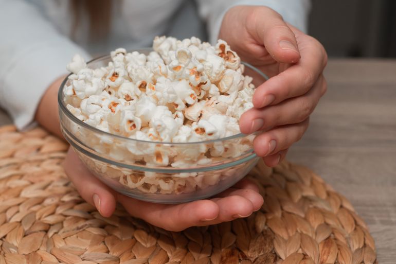 pop2-1730390354 Woman Holding Bowl of Fresh Popcorn. Close-up of a woman's hands holding a transparent bowl filled with freshly popped popcorn, on a natural woven mat