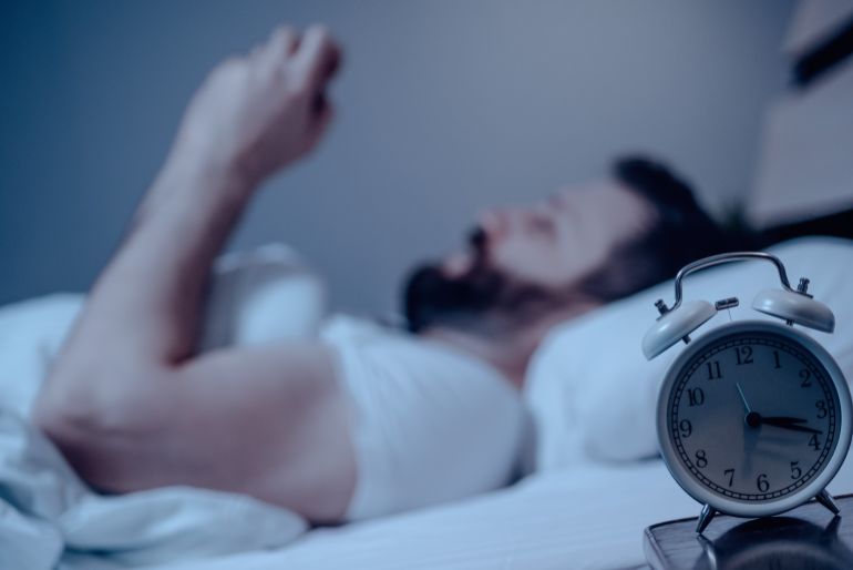 Alarm clock on the bedside table. A lonely bearded dark-haired man lies on the bed at night and is chatting or playing on the phone. Close-up, insomnia, bedroom, self-isolation, top view.One