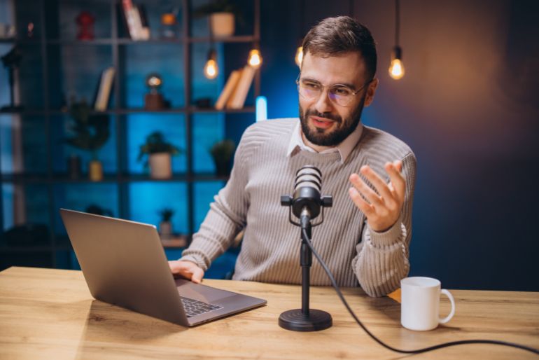 Podcaster wearing headphones, speaking into condenser microphone while working on laptop at home workspace