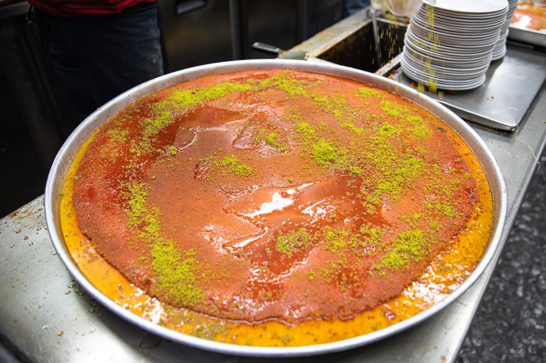 'Kanafeh Nabulsiyeh', a traditional Middle Eastern dessert made with fine semolina dough, soaked in sweet, sugar-based syrup, and typically layered with cheese topped with crushed pistachio nuts ; Shutterstock ID 1700732473; purchase_order: aj; job: ; client: ; other:
