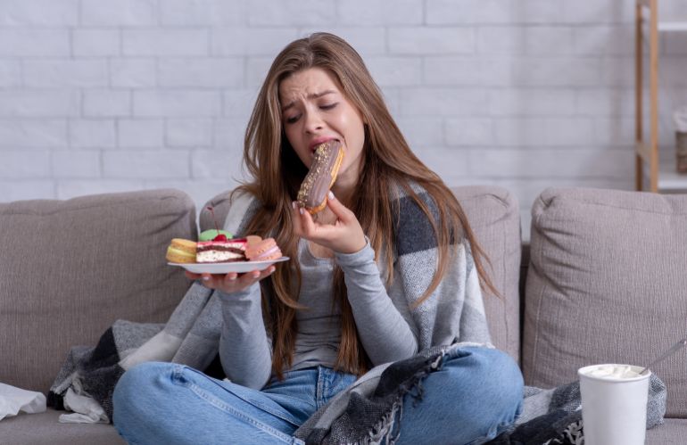 Comfort foods and stress eating concept. Frustrated young lady devouring pastry on sofa at home. Upset woman dealing with negative emotions, anxiety or problems through sweets; Shutterstock ID 1836395869; purchase_order: ajnet; job: ; client: ; other: