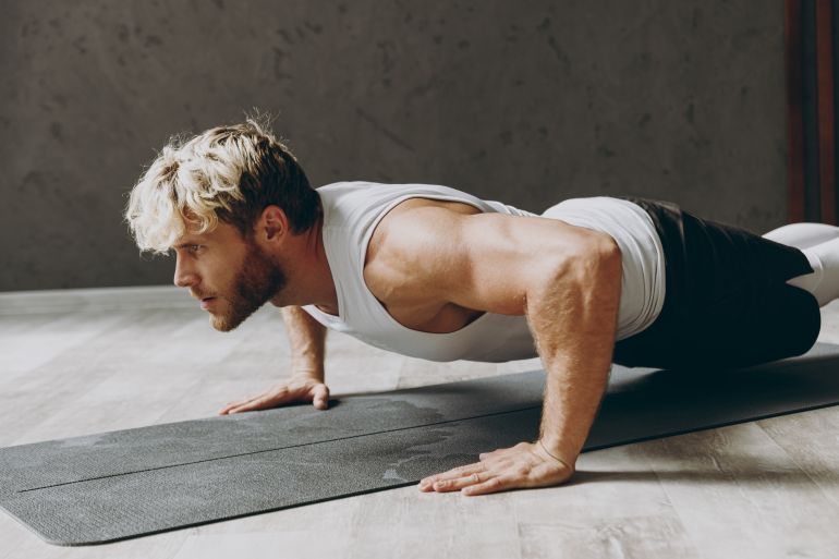 Side profile view calm confident young strong sporty athletic sportsman man wear white tank shirt black shorts doing push-ups exercises on floor warm up training indoor at gym. Workout sport concept