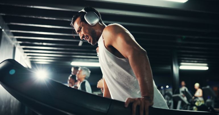Man, treadmill and tired with sweat at gym with exercise, headphones and speed with break at training center. Person, running machine and fatigue with workout, breathing and burnout at fitness club