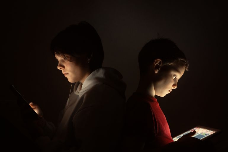 tr6gy-1770619809 Brother and sister use smartphone and digital tablet while sitting back to back at night at home. Childhood addiction to digital mobile devices