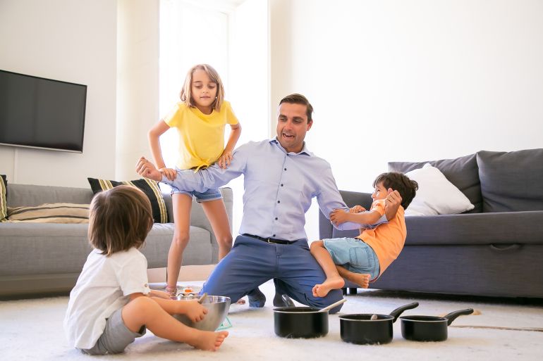 091-1-1773140001 Happy Caucasian man playing with children and showing strength. Cheerful kids having fun together in living room on carpet. Pans and bowl for game. Childhood, weekend and home activity concept في بيك