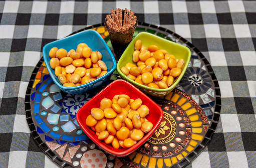 d8aad8b1d985d8b3-1774098176 Top view. On the table with a checkered tablecloth we see a mosaic sideboard with three small bowls of lupin beans ready to eat. Above on the sideboard we see an antique silver toothpick holder with many wooden toothpicks. Lupin beans are very popular on any occasion. In the tropical autumn. São Paulo, São Paulo state, Brazil.