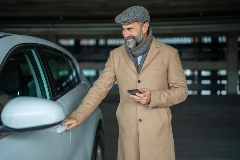 Mature man with a stylish coat and cap smiling while opening a car door in an indoor parking garage, holding a mobile phone and preparing for his urban commute