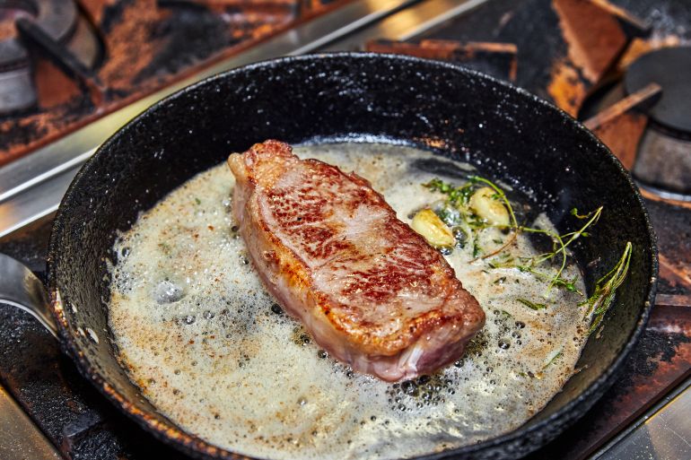 Frying pan on a stove on which a large piece of meat is fried with butter