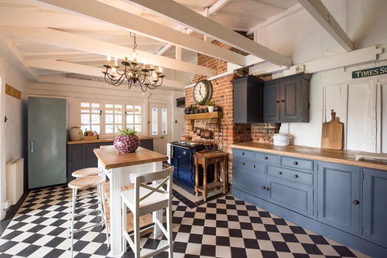 A general interior view of an old English country cottage style kitchen, with black and white tiled floor, painted dining table with stained wood top with chairs and bar stools, blue cabinets, chandelier, bare brick chimney breast with Aga range cooker, large clock, chunky wooden mantel shelf and exposed ceiling roof timber beams