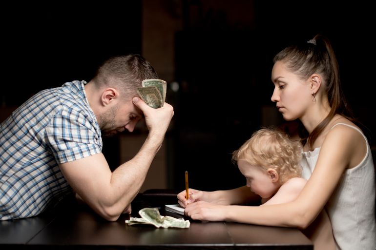 shutterstock_1764571841-1774271307 A family with a small child considers modest monetary savings late in the evening. Father sadly counts dollars, mother and little child are sitting opposite. The financial crisis in a young family
