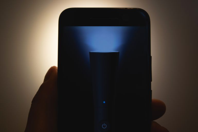 The hand holds a smart phone with the flashlight on in the dark.