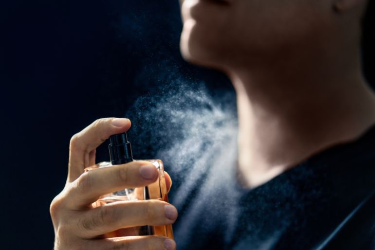 shutterstock_2383455475-1774608199 Fragrance spray. Man and perfume. Applying cologne. Scent water. Skin care, beauty product and male cosmetics concept. Holding elegant smell bottle in hand. Dark blue background. Young gentleman.