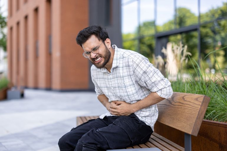 shutterstock_2511048691-1773934230 Contorted from severe pain, a young Indian man sits on a bench in the street and holds his stomach with his hands.; Shutterstock ID 2511048691; purchase_order: aj; job: ; client: ; other:
