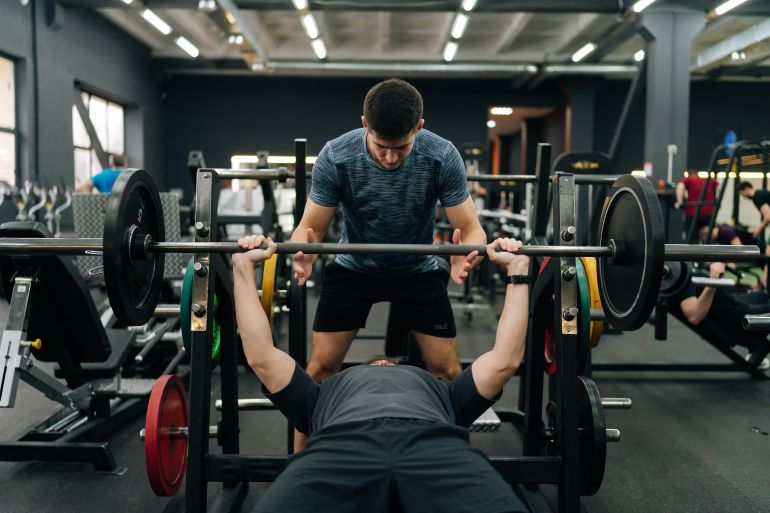 Ufa, Russia - 27 March 2024. Portrait of personal fitness trainer helping beginner sportsman doing barbell bench press exercise during workout in gym. Athletic male doing barbell bench press under