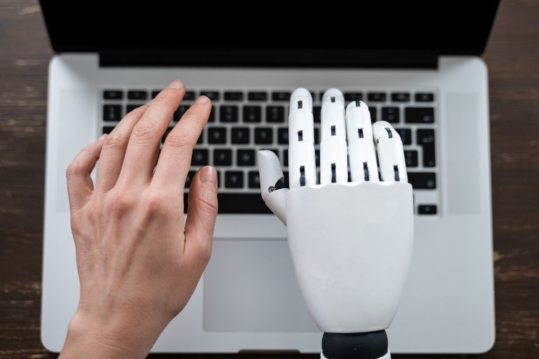 Human and robot hands positioned above laptop keyboard, AI and human collaboration in coding, writing and digital tasks