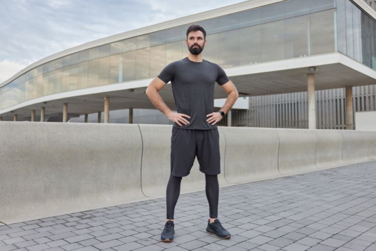 Horizontal shot of sporty man has regular training outdoors being in good physical shape has muscular arms dressed in sportswear keeps hands on waist maintains healthy lifestyle poses in urban setting