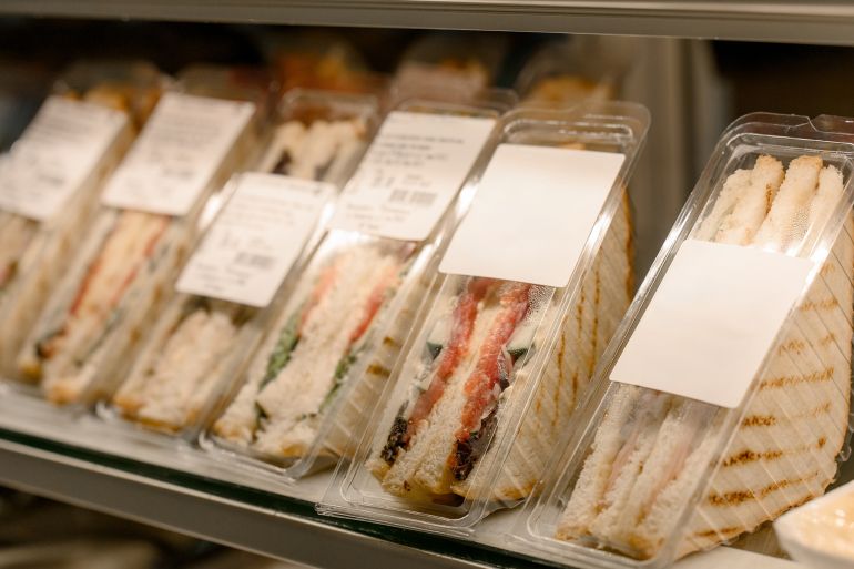 A row of triangular sandwiches in clear plastic packaging on a store display shelf. Close-up of ready-made sandwiches and club sandwiches with ham, cheese and vegetables in packaging