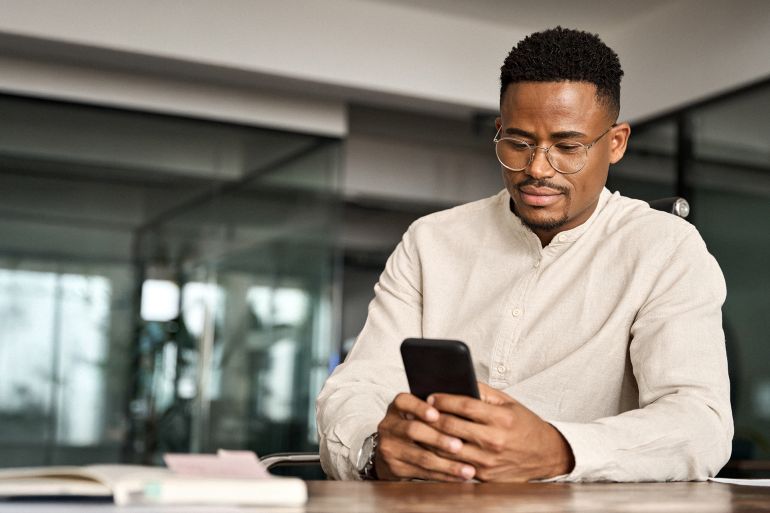Busy African American entrepreneur using cell phone working in office. Professional young Black business man holding mobile cellphone looking at cellphone making financial bank payment at work.