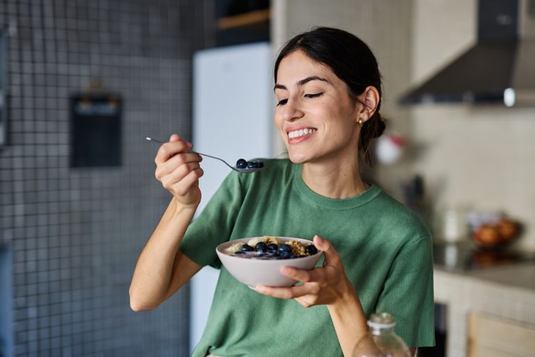 64984sd-1770038073 Portrait of a young woman preparing and eating breakfast, drinking juice in the kitchen at home
