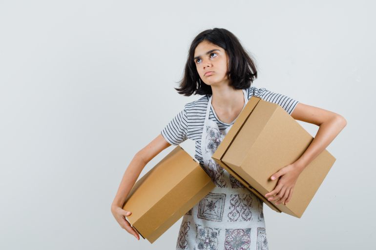 Little girl holding cardboard boxes in t-shirt, apron and looking pensive , front view. freepik