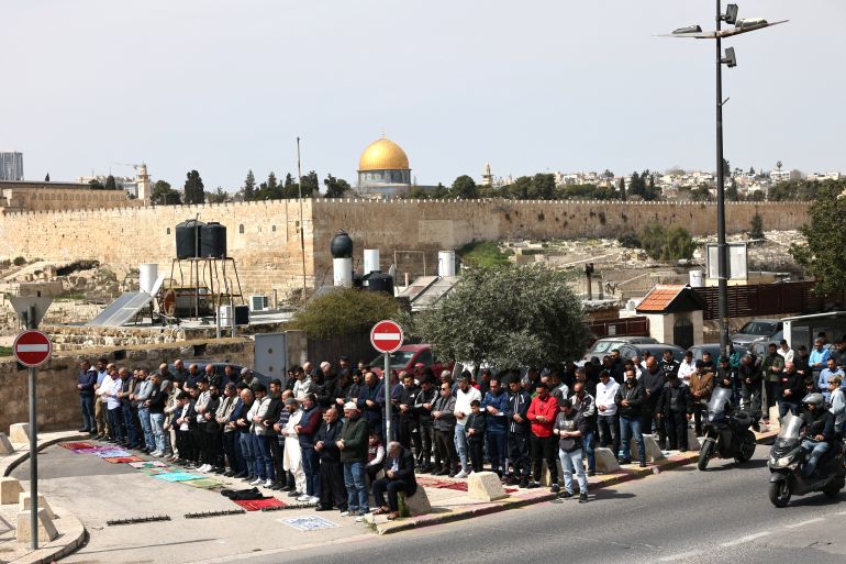 Palestinian Muslims pray during the last Friday noon prayer of the Muslim holy fasting month of Ramadan, on a street blocked by Israeli security forces in the east Jerusalem neighbourhood of Ras al-Amud, on March 13, 2026, as the site al-Aqsa mosque and Dome of the Rock (background) is closed since the US-Israel war with Iran started on February 28. (Photo by AHMAD GHARABLI / AFP)