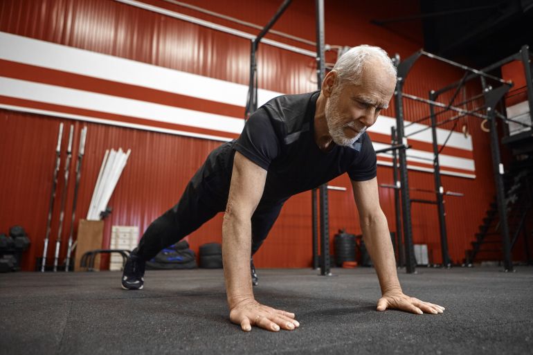 Healthcare, age, retirement and rehabilitation concept. Muscular fit seventy year old unshaven man in sportswear doing plank in gym. Senior male planking during morning workout at fitness center