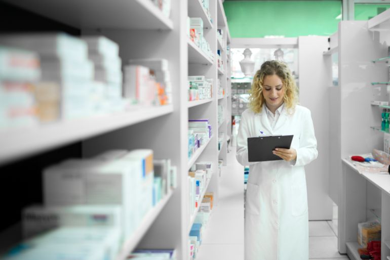 Pharmacist in white uniform walking by the shelf with medicines and checking inventory.