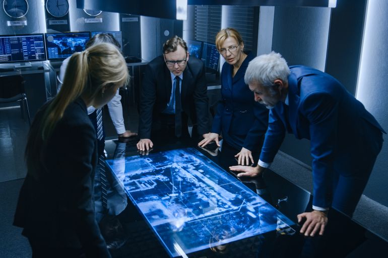 Team of Government Intelligence / FBI Agents Standing Around Digital Touch Screen Table and Tracking Suspect Vehicle Using Satellite Surveillance in the Monitoring Room.