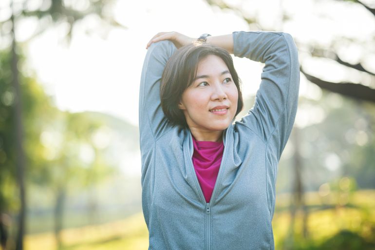Warm Up and Cool Down Stretching Exercises. Asian female runner in sportswear doing overhead triceps stretch before morning jogging on trails in public park.