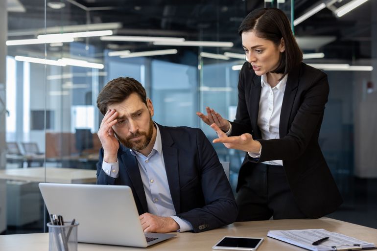 Conflicted office scene showing an angry female boss yelling at a frustrated male employee working on laptop. Tension highlights workplace stress and pressure during a business meeting.