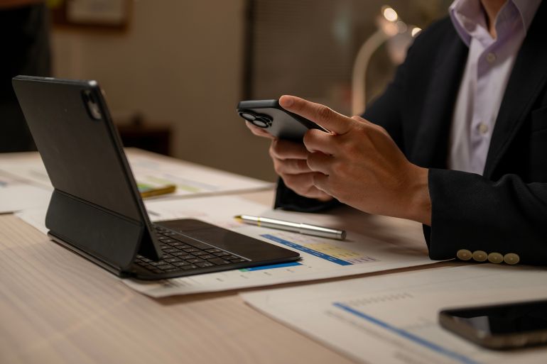 A man is using a cell phone while sitting at a desk. There are two cell phones on the desk, one of which is being used by the man