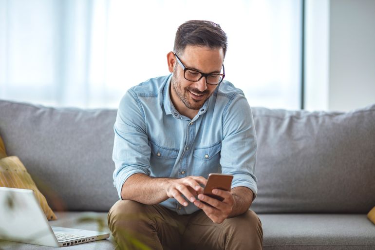 Man with spectacles relaxing sitting on couch while looking at mobile phone. Closeup of mature man using smartphone to checking email at home. Man reading email on smartphone; Shutterstock ID 1767975917; purchase_order: aj; job: ; client: ; other: