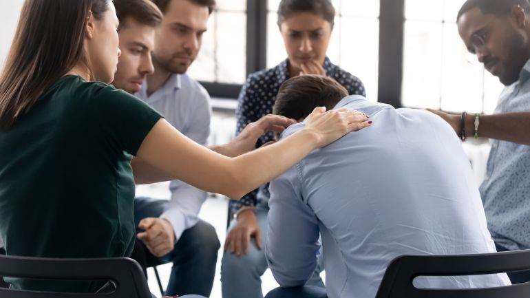 Rear view diverse people comforting frustrated depressed man at meeting, helping overcoming problem, drug or alcohol addiction, sitting in circle at group counselling session, mental healthcare