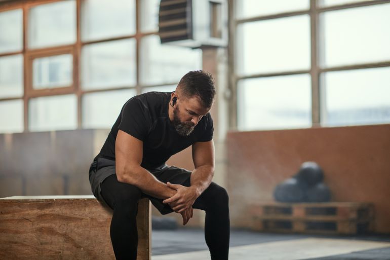 Strong bearded male athlete in wireless earphones sitting on box and listening to music during break in intense training in gym