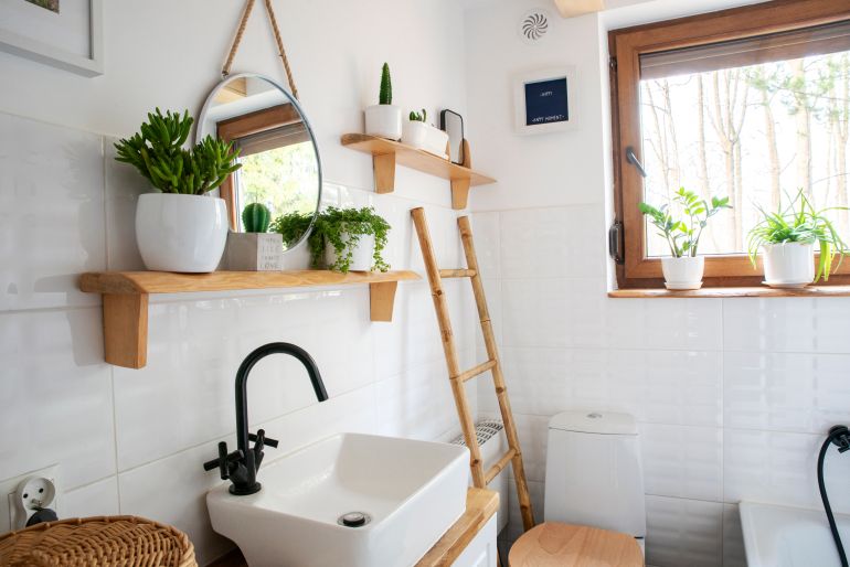 Interior of small bathroom with window, wooden decoration, shelf and ladder. Stylish wash basin and tap, round mirror on a white wall and plants on a shelf. Rustic bathroom in a cottage in rural style FW: تقرير عن ترتيب المنزل الضيق