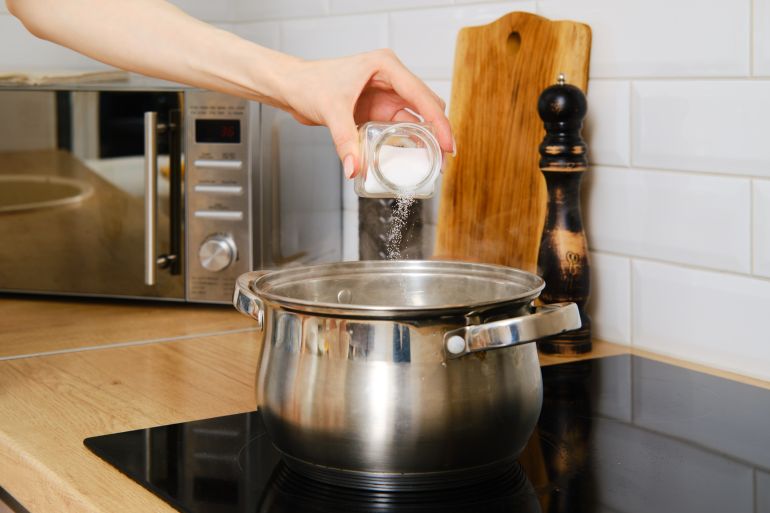Closeup view of female hand pouring salt to water boiling on electric stove. Hand with salt over stainless steel stockpot.