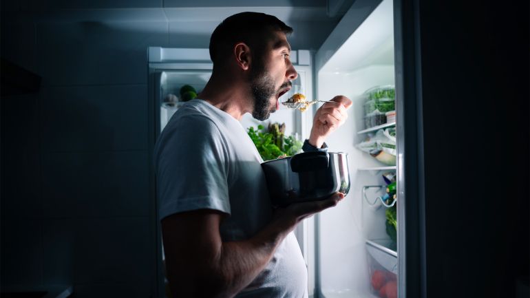 Hungry man eating food at night from open fridge. Man taking midnight snack from refrigerator;:
