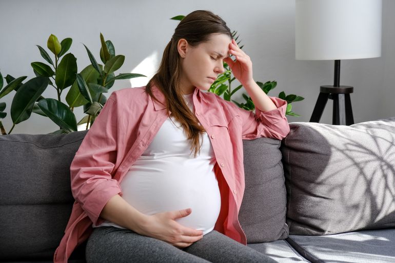 shutterstock_2273460889-1767119443 Tired sad young pregnant female suffering migraine sitting alone on sofa at home. Unhappy future mom holding painful head while having headache and frowning, sickness woman touching big belly; Shutterstock ID 2273460889; purchase_order: aj; job: ; client: ; other: